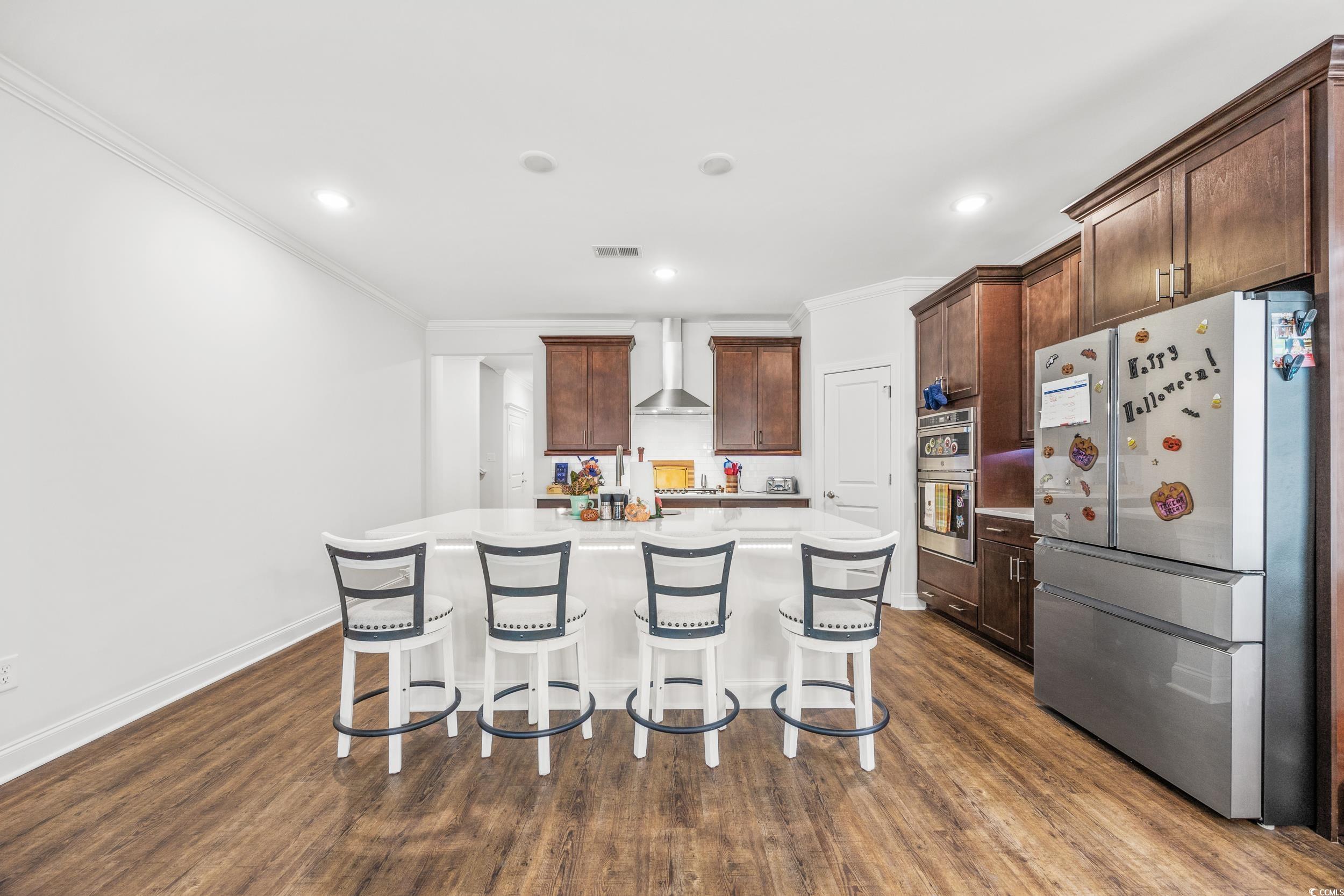 Kitchen with wall chimney range hood, a center island with sink, a kitchen bar, dark hardwood / wood-style floors, and stainless steel appliances