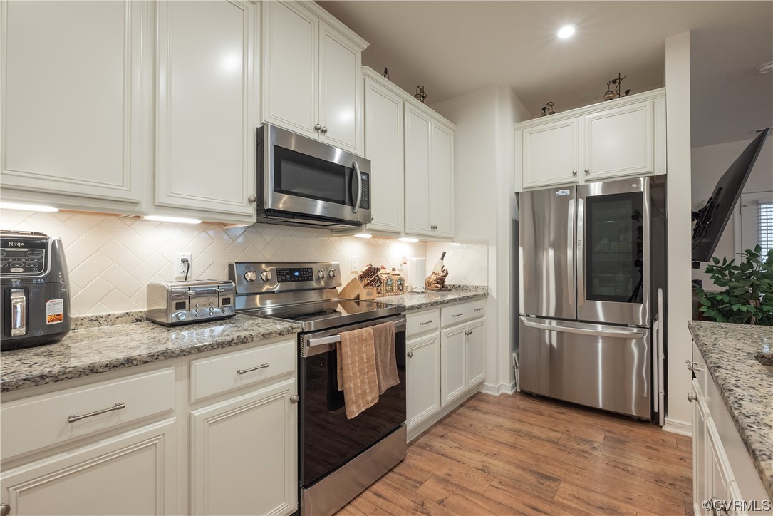 Kitchen with white cabinets, light hardwood / wood-style flooring, stainless steel appliances, light stone counters, and backsplash