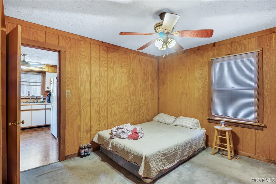 Carpeted bedroom featuring a textured ceiling, white fridge, sink, wooden walls, and ceiling fan