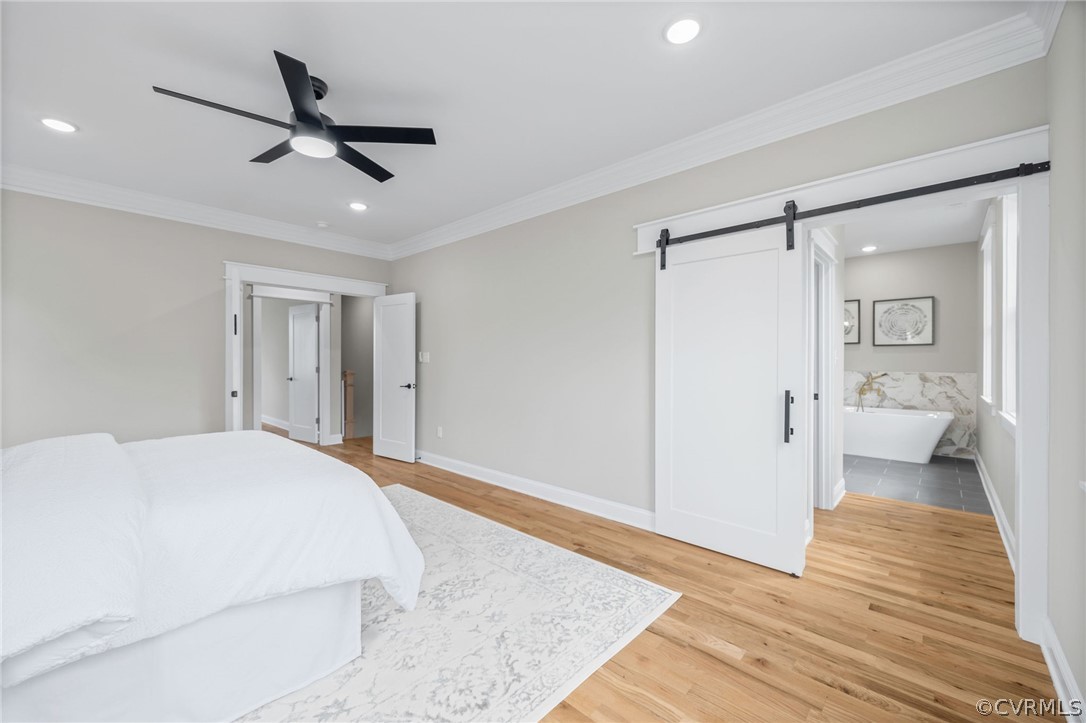 Bedroom with a barn door, ceiling fan, light wood-type flooring, connected bathroom, and ornamental molding