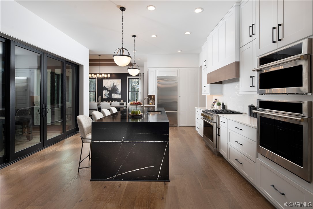 Kitchen featuring premium appliances, hanging light fixtures, wood-type flooring, and an island with sink