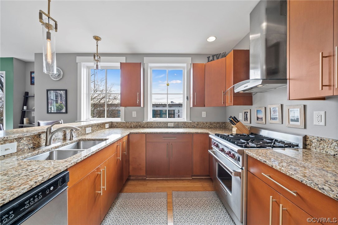 Kitchen featuring wall chimney range hood, stainless steel dishwasher, light hardwood / wood-style floors, light stone counters, and gas range oven