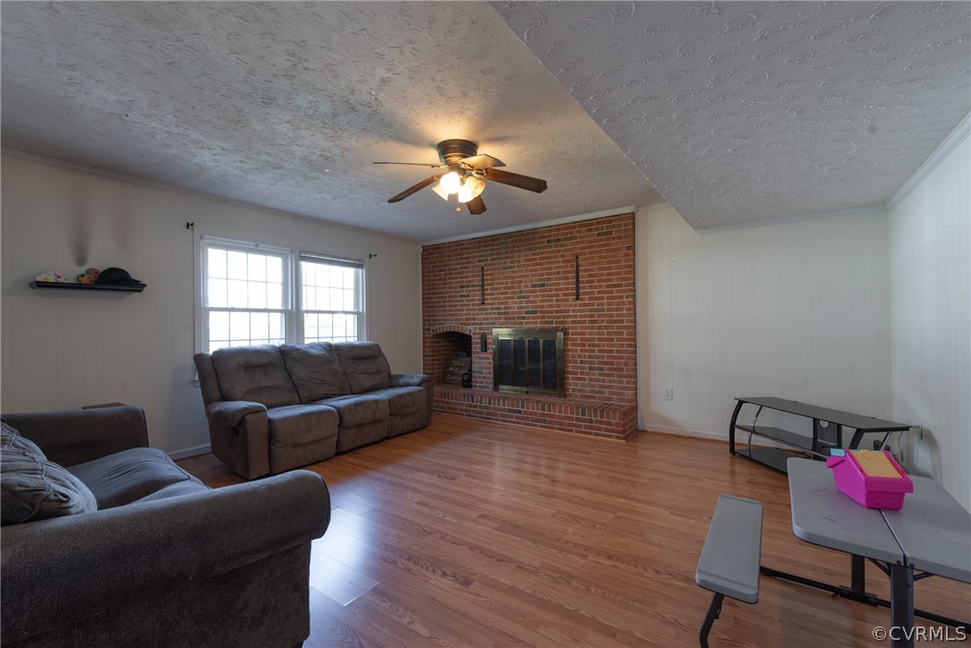 Living room with a textured ceiling, a brick fireplace, ornamental molding, wood-type flooring, and ceiling fan
