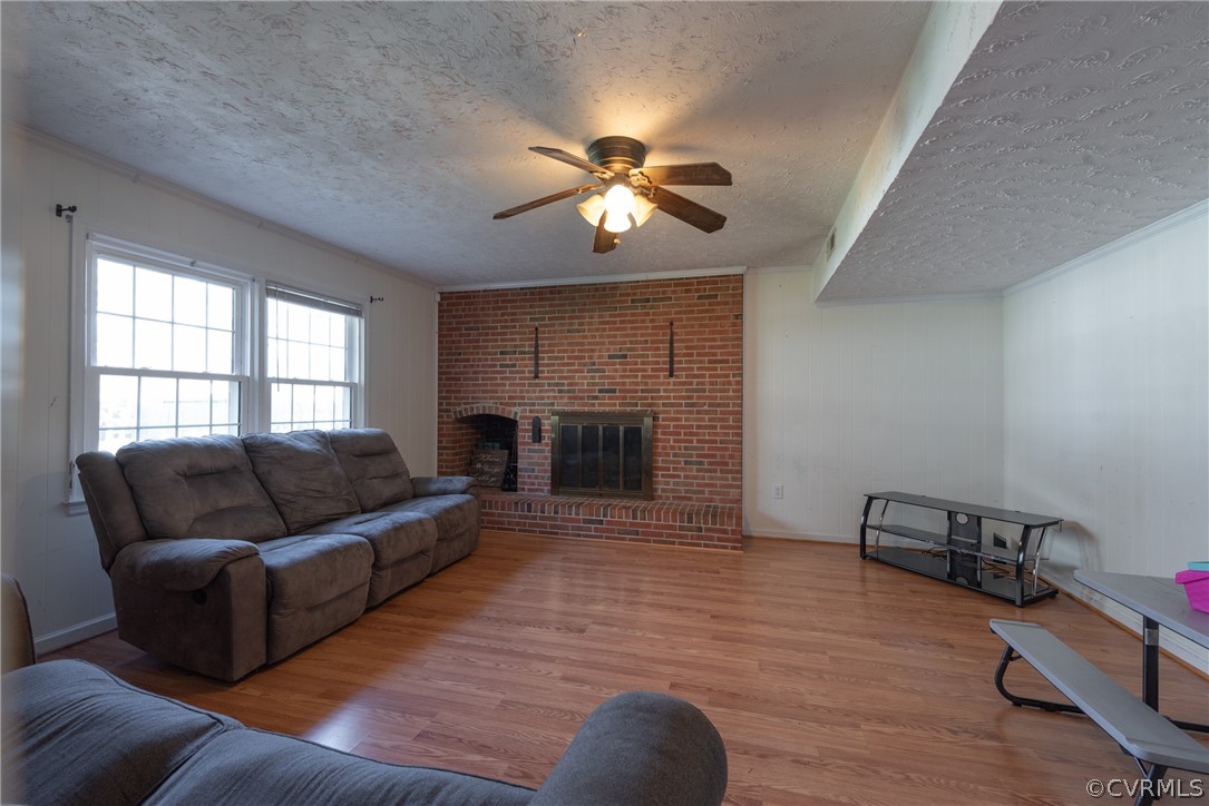 Living room featuring a textured ceiling, a brick fireplace, ceiling fan, light hardwood / wood-style flooring, and crown molding