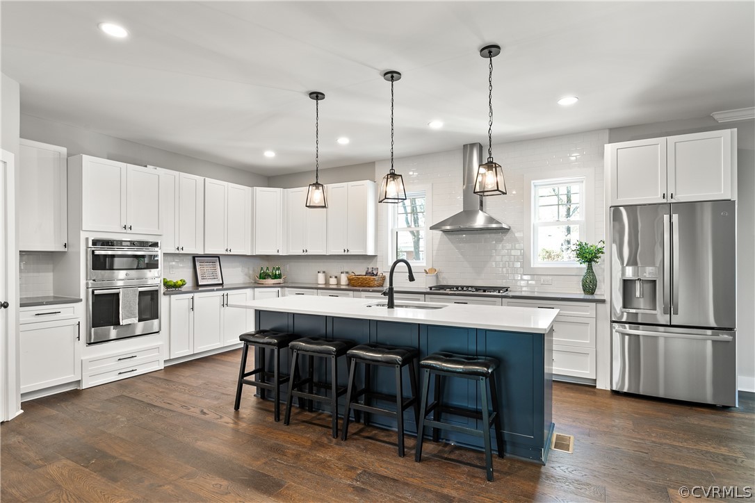 Kitchen featuring stainless steel appliances, dark hardwood / wood-style floors, wall chimney exhaust hood, a kitchen island with sink, and sink