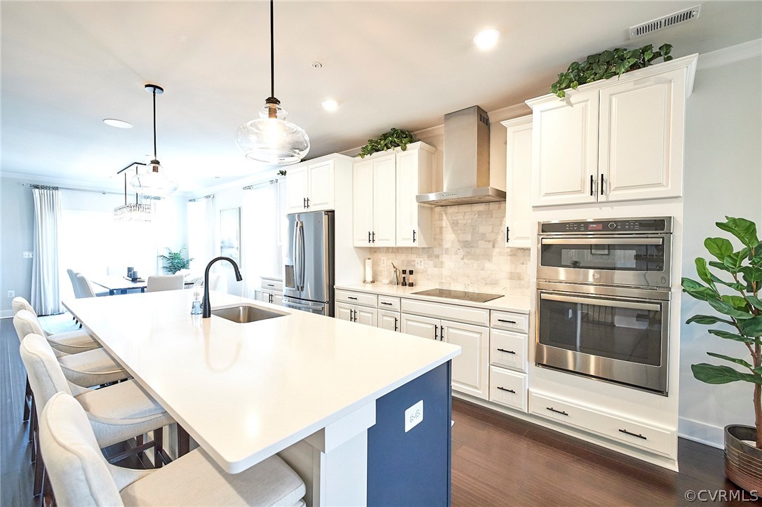 Kitchen featuring a kitchen bar, wall chimney range hood, decorative light fixtures, stainless steel appliances, and a center island with sink