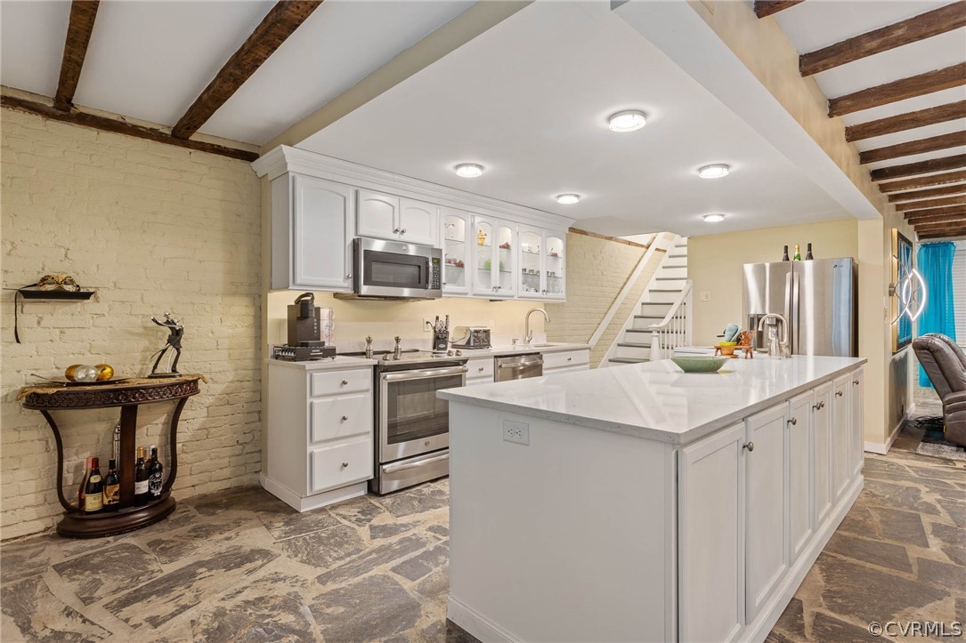 Kitchen featuring beamed ceiling, white cabinetry, brick wall, appliances with stainless steel finishes, and an island with sink