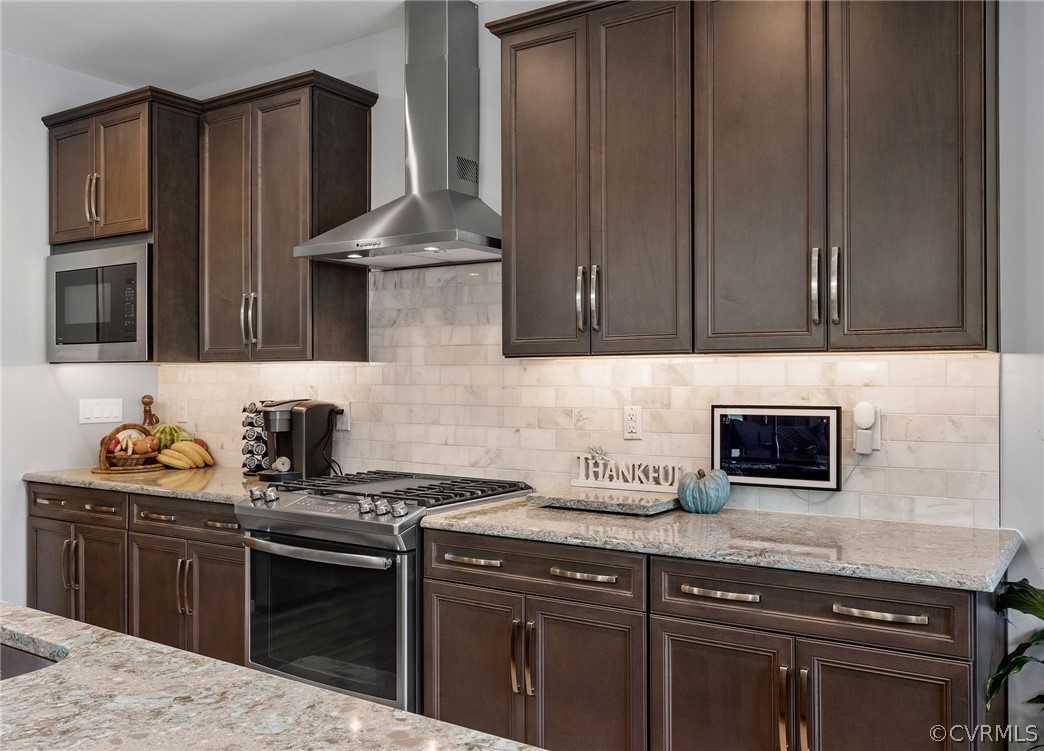 Kitchen with range with gas stovetop, light stone countertops, stainless steel microwave, tasteful backsplash, and wall chimney exhaust hood