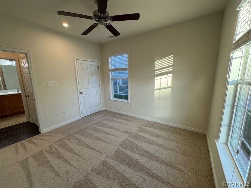 Unfurnished bedroom with ensuite bath, light colored carpet, ceiling fan, and multiple windows
