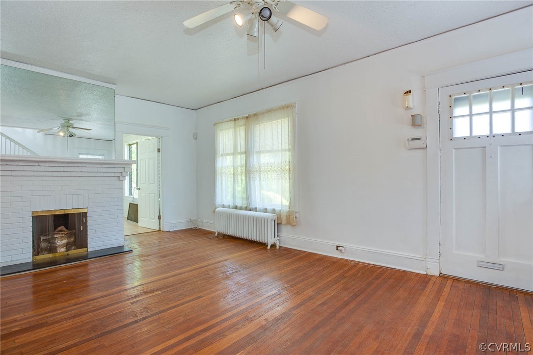 Unfurnished living room featuring wood-type flooring, ceiling fan, a brick fireplace, and radiator