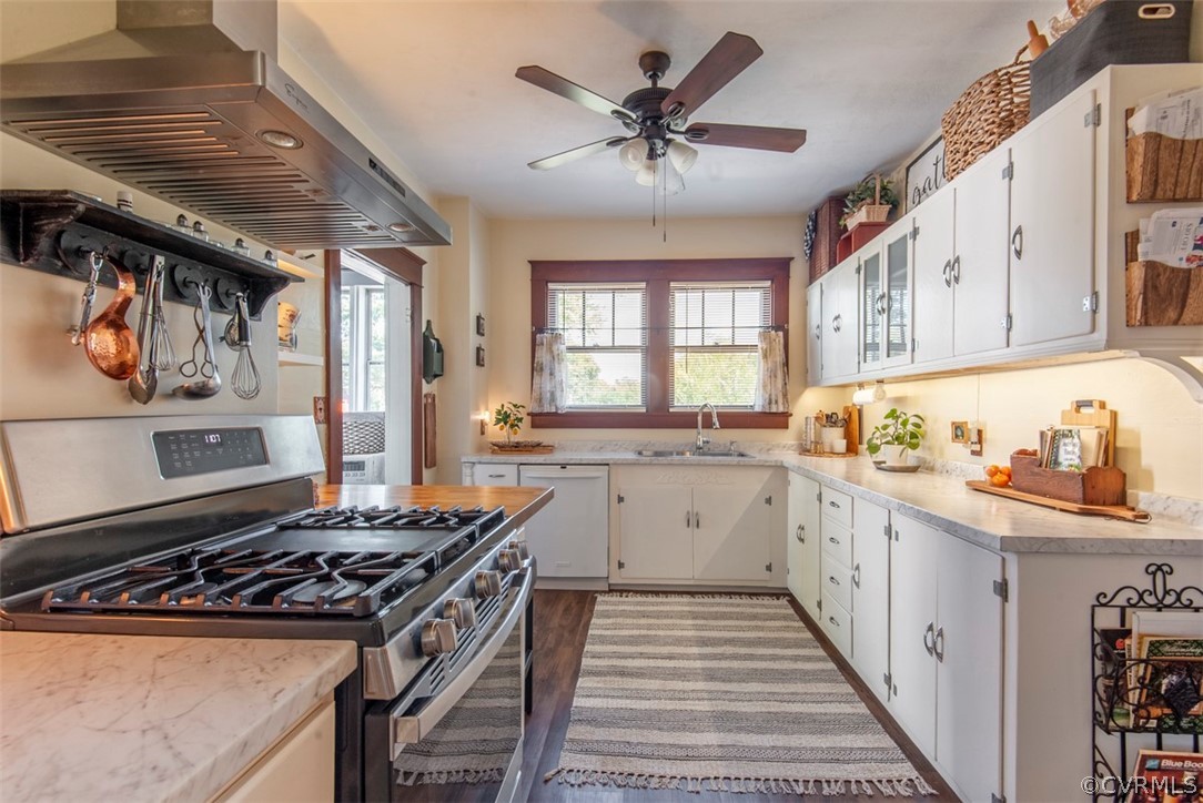 Kitchen featuring ceiling fan, white dishwasher, wall chimney exhaust hood, white cabinetry, and stainless steel gas range