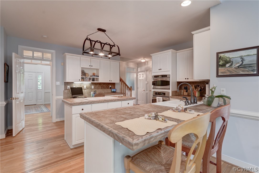 Kitchen featuring double oven, white cabinets, tasteful backsplash, hanging light fixtures, and light hardwood  flooring