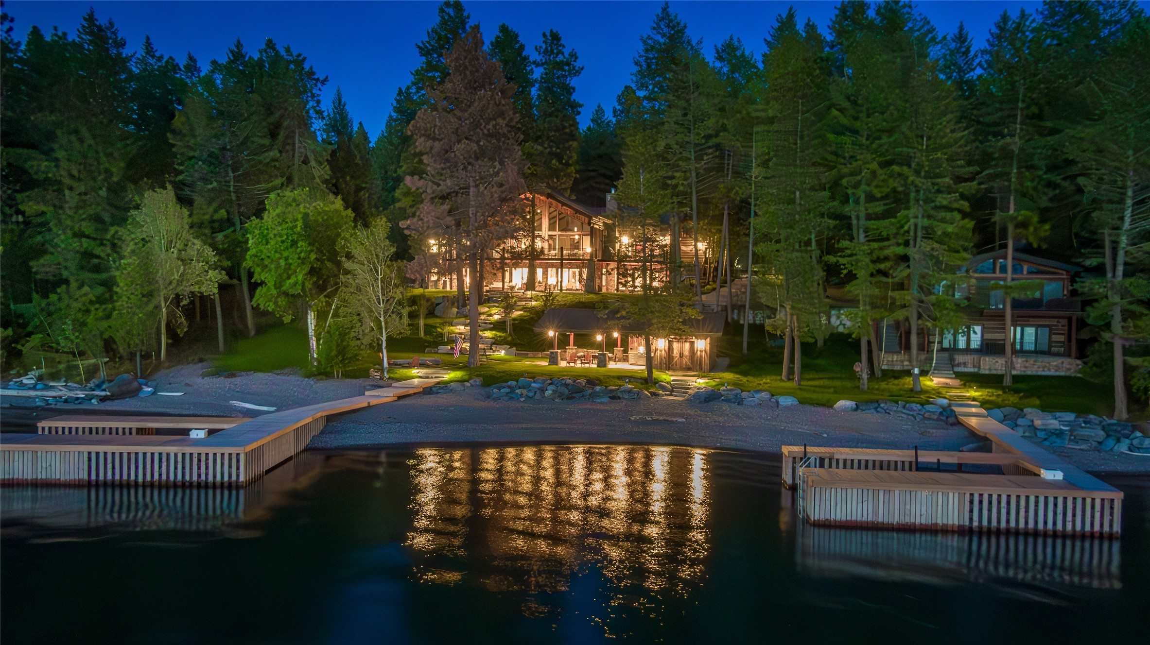 Indulge in the enchanting allure of this extraordinary property, gracing the shores of Flathead Lake with a remarkable 208 feet of prime waterfront. Situated just minutes away from the charming Village of Bigfork, this unique estate boasts two extensively remodeled homes, each meticulously crafted to showcase unparalleled attention to detail, each home on their own tract of land. Upon entering the main home, the expansive main floor reveals an awe-inspiring view of Flathead Lake that treats you to its legendary sunsets. The living room captivates with a majestic rock fireplace and lofty exposed beams, seamlessly transitioning to a sprawling covered deck where panoramic views become your daily backdrop. The kitchen stands as a masterpiece of design, adorned with custom cabinetry and a stunning wall of storage, creating a haven for culinary enthusiasts and making entertaining a true delight. The main floor bedroom suite offers a retreat of luxury, featuring separate walk-in closets, dual vanities, a spacious tile shower, and a soaking tub strategically positioned to overlook the beautiful landscape and Flathead Lake. For those who value a dedicated workspace, the office/TV room beckons with built-in desks for two, providing a cozy spot for fireside relaxation and convenient access outside through patio doors. A generously sized laundry/storage room effortlessly connects to the 3-stall garage and additional storage space. The lower level of the home unfolds into an expansive entertainment area, characterized by high ceilings, another inviting fireplace, and a convenient wet bar. Double doors lead to the outdoor patio, a perfect setting for gatherings, creating a seamless transition between indoor and outdoor living. This property transcends the definition of a home; it is a gateway to lakeside living at its finest, incredible landscaping with large stone stairs to the waters edge. Also boasting spacious guest house and at the lake level – an outdoor living space complete with kitchen, entertaining and bathroom. Every detail has been thoughtfully curated for your comfort and enjoyment, offering an unparalleled opportunity to savor the beauty of Flathead Lake in a setting where luxury and natural splendor harmoniously coexist.  Main home has a total of 7 bedrooms, non-conforming, septic is built for 3 bedrooms; guest house can sleep 8, septic is built for 2 bedrooms.  Sellers are 2 with occasional guests.  Welcome to a residence where each moment is an invitation to embrace the art of. For your private showing call Cherie T. Hansen 406-253-4546 or your real estate professional.
