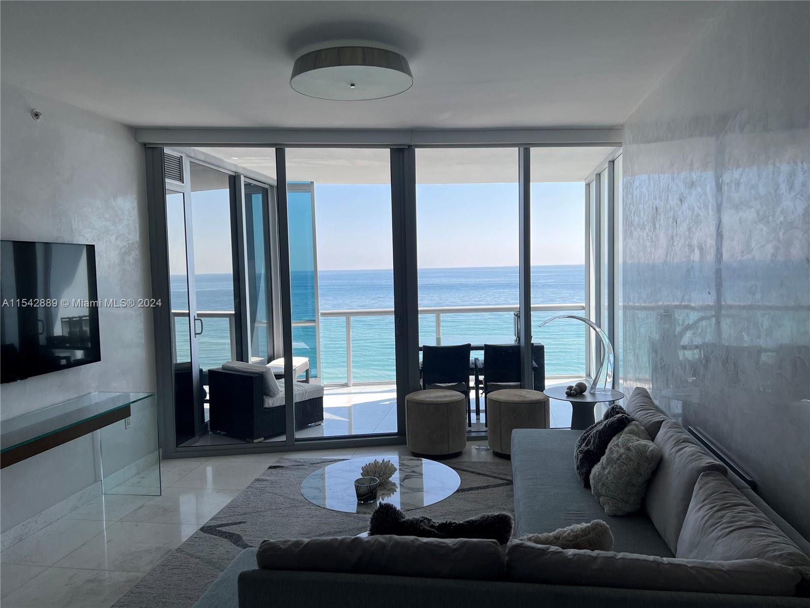 As you enter this apt, your eyes are immediately drawn to the stunning panoramic view of the sea. The floor-to-ceiling windows allow natural light to flood the room, illuminating the elegant décor providing a sense of spaciousness. Painted in pearl grey, creates a calming atmosphere which the L-shaped sofa with soft pillows invites you to sit and relax . A round marble coffee table sits in the center of the room, adding a touch of sophistication A crystal media console is situated against one wall, framing the base of a flat-screen TV. Windows are dressed with motorized sheer white sunscreens, casting a warm and inviting glow over the room. Overall, whether lounging pool side or overlooking the sea, this condo offers a luxurious and comfortable retreat, a perfect place to call home.