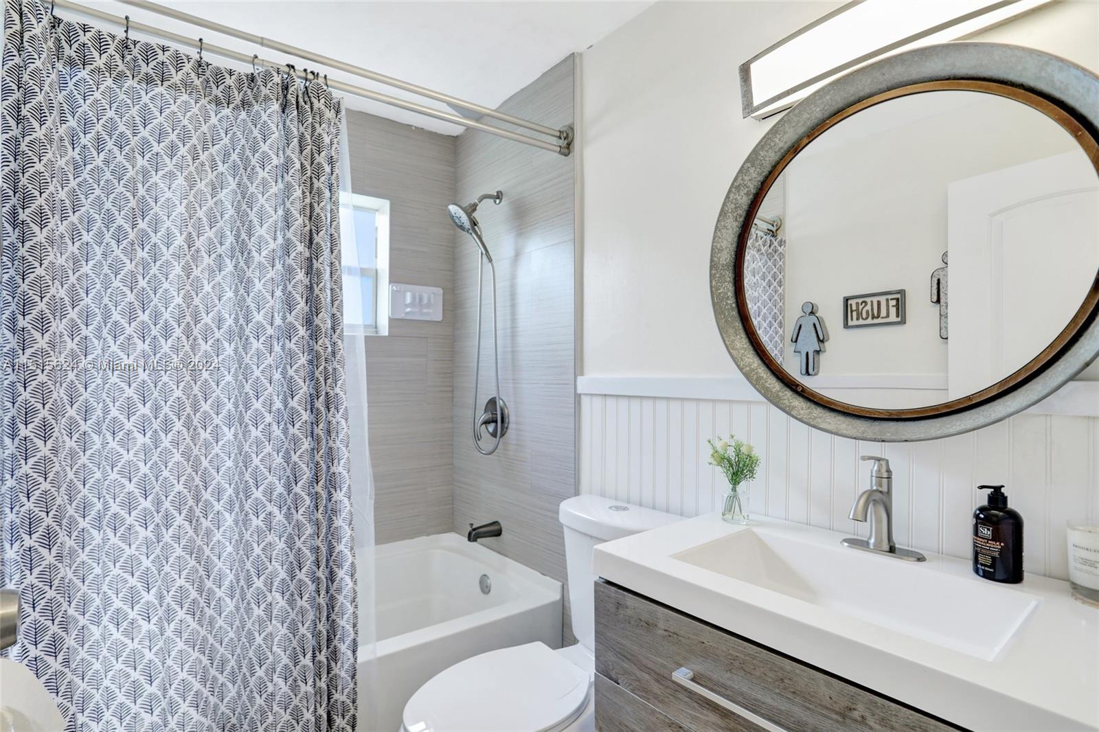 This serene bathroom blends cool, contemporary lines with rustic textures. A leaf-patterned shower curtain adds a touch of nature, while the circular mirror reflects a well-lit, tidy space that invites a refreshing start or a relaxing end to any day.
